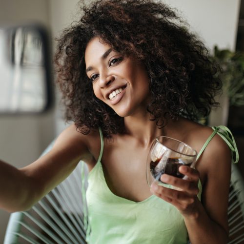 African woman in silk green top takes selfie and holds coffee glass. Dark-skinned curly lady smiles