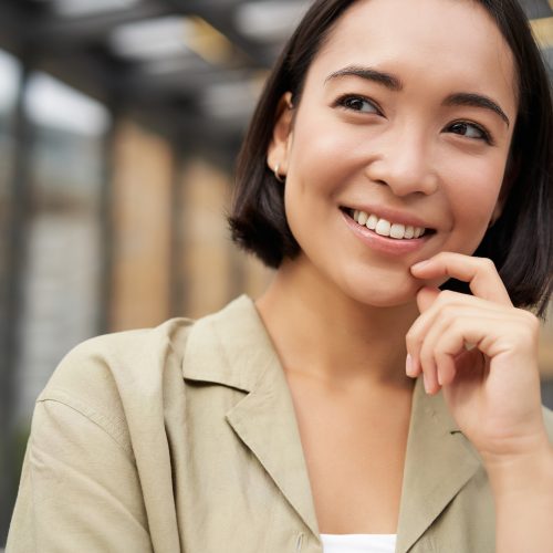 Close up portrait of asian girl with happy face, smiling and laughing, touching her skin, standing