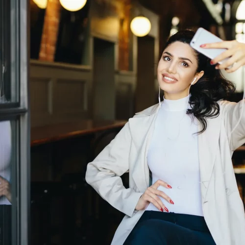 Woman with dark hair tied in pony tail wearing white jacket sitting in cafe making selfie of her eyelash extensions.