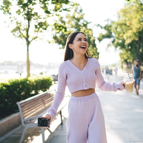 Millennial with smartphone and coffee laughing in city street